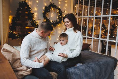 Yürek ısıtan bir sahnede, samimi süveter giyen sevgi dolu ebeveynler, parıldayan ışıklar ve şenlikli dekorasyonlarla çevrili neşeli çocuklarıyla vakit geçirip, sıcaklık yaratıyorlar.