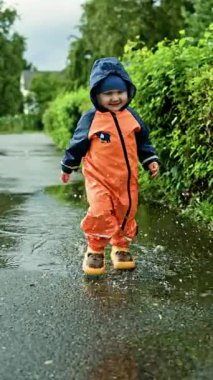 Çocuk lastik botlar ve su geçirmez tulumlarla su birikintilerinde koşuyor. Çocuk eğleniyor ve yağmur sırasında su birikintilerinde koşuyor. Neşeli bir çocukluk. Çocuk yağmurluğu..