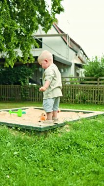 Kum havuzunda oynayan, tişörtlü ve şortlu sarışın 2 yaşında bir çocuk. Çocuk parkta kum kazıyor. Çocuk dışarıda oynuyor. Erkekler için şık yaz kıyafetleri..