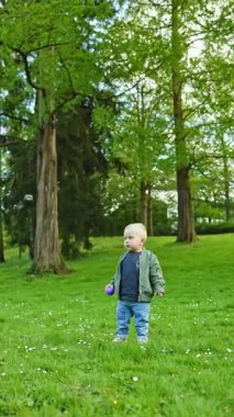 Neşeli çocuk şehir parkında çimlerin üzerinde yürüyor. Çocuk temiz havada yürüyüşe çıkmış. Ceketli ve pantolonlu bir çocuk yeşil çimlerde yürüyor. Çocuk gelişimi.
