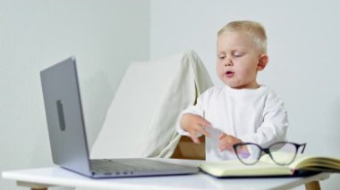 2 yaşında, gözlüklü, dizüstü bilgisayarlı, evde masada oturmuş, online aktivitelere odaklanmış bir çocuk. Teknoloji ve çocukluk kavramı.
