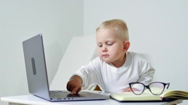 Gözlüklü küçük 2 yaşında bir çocuk bir iş adamı görüntüsünde dizüstü bilgisayarın başında oturuyor. Çocuk çok meşgul ve internette çalışıyor. Çocuk bir ofis çalışanı..