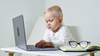 Gözlüklü akıllı 2 yaşındaki çocuk evdeki masada dizüstü bilgisayar kullanıyor. Çocuk uzaktan öğrenme sırasında bilgisayarın başında oturur. Çocukluk ve modern teknolojiler.