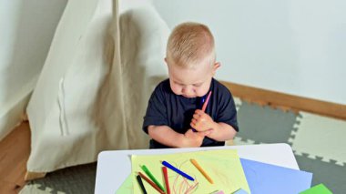 Küçük bir çocuk, mavi tişörtlü bir bebek, bir masaya oturur ve kalemlerle resim çizer. Çocuğun önünde dağınık renkli kalemler var. Çocuk eline bir kalem alıp kağıda çiziyor..