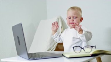 Gözlüklü küçük 2 yaşında bir çocuk bir iş adamı görüntüsünde dizüstü bilgisayarın başında oturuyor. Çocuk çok meşgul ve internette çalışıyor. Çocuk bir ofis çalışanı..