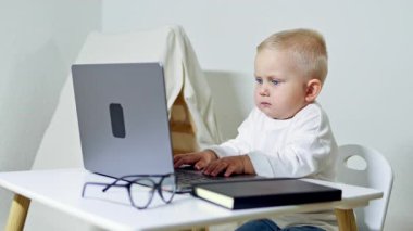 Gözlüklü küçük 2 yaşında bir çocuk bir iş adamı görüntüsünde dizüstü bilgisayarın başında oturuyor. Çocuk çok meşgul ve internette çalışıyor. Çocuk bir ofis çalışanı..