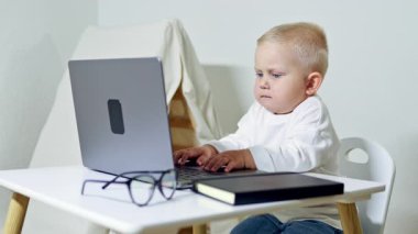 Gözlüklü akıllı 2 yaşındaki çocuk evdeki masada dizüstü bilgisayar kullanıyor. Çocuk uzaktan öğrenme sırasında bilgisayarın başında oturur. Çocukluk ve modern teknolojiler.