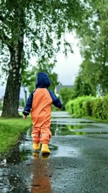 2 yaşında, su geçirmez tulumlu ve lastik çizmeli bir çocuk. Çocuk ve yağmur sırasında su birikintilerinde koşmak. Neşeli tasasız bir çocukluk. Çocuklar için su geçirmez giysi.
