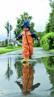 Su geçirmez tulumlu neşeli 2 yaşında bir çocuk. Yağmurluklu ve lastik botlu çocuk su birikintilerinde koşuyor. Oğlan sokakta yağmurda koşuyor.