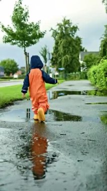 Çocuk lastik botlar ve su geçirmez tulumlarla su birikintilerinde koşuyor. Çocuk eğleniyor ve yağmur sırasında su birikintilerinde koşuyor. Neşeli bir çocukluk. Çocuk yağmurluğu..