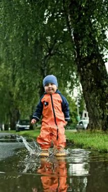 Parktaki yağmur sırasında büyük bir su birikintisinde koşan ve oynayan lastik çizmeli mutlu küçük çocuğun videosu. Su geçirmez tulumla su birikintilerinde koşan bir bebek. Çocuk yağmurda eğleniyor..