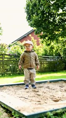 Oyun parkındaki kum havuzunda kepli şirin iki yaşında bir çocuk oynuyor. Şehir parkında elinde kum havuzuyla gezen şık elbiseli bir çocuk. Çocuk kumu kazıyor ve sokakta oynuyor..