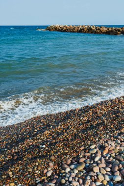 Çeşitli büyüklükteki çakıl taşları, parlak, güneşli bir öğleden sonra açık mavi gökyüzünün altında, kayalık bir iskelenin yakınında huzurlu bir manzara oluşturan hafif dalgalar gibi kıyıları kaplıyor..