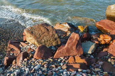 Dalgalar renkli kaya ve çakıl taşlarına çarpar manzaralı bir kıyı şeridi boyunca, güneşin altında canlı bir sahil gününün özünü yakalar..