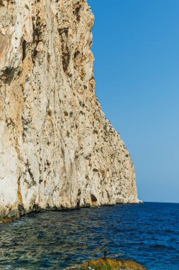 Görkemli kayalıklar bulutsuz bir gökyüzünün altında canlı denizle buluşarak çarpıcı bir manzara yaratıyor. Sakin su doğanın güzelliğini, keşif ve macerayı yansıtır..