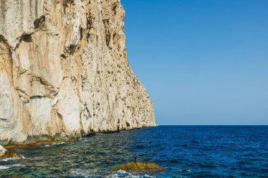 Güneş ışığının kayalık yüzeyleri aydınlattığı görkemli uçurumlar parlak mavi sulardan keskin bir şekilde yükselir. Durgun deniz, parlak ve bulutsuz bir gökyüzünün altında sonsuza dek uzanır..