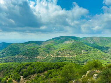 Yuvarlanan yeşil tepeler, parçalı bulutlu bir gökyüzünün altında uzanıyor. Gün boyunca huzurlu bir dağlık bölgede doğanın güzelliğini sergiliyor..