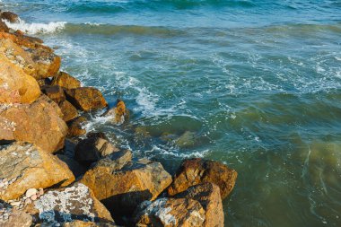Güneş batarken, dalgalar yumuşak bir şekilde kayalık bir kıyıda süzülüyor ve suya sıcak bir parıltı saçıyor. Huzurlu sahne, okyanusun rahatlama ve yansımasını davet eder..