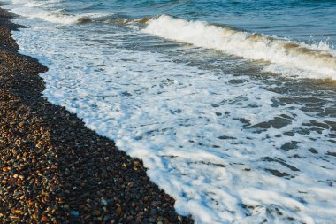 Sabahın erken saatlerinde kıyı şeridini aydınlatan hafif dalgalar renkli çakıl taşlarına karşı beyaz köpük oluşturur. Sahne huzuru ve doğayla bağlantıyı çağrıştırıyor..