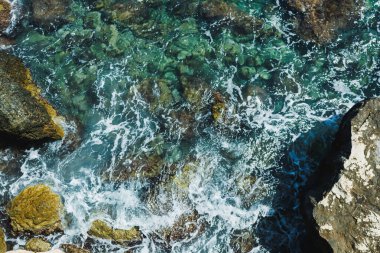 Yukarıdan çekilen temiz turkuaz dalgalar engebeli kayaların etrafında köpük saçıyor. Güneş ışığı suda parlıyor, rahatlatıcı ve güzel bir sahil anı yaratıyor..