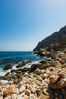 Parlak güneş, kayalık bir kıyı şeridiyle huzurlu bir plajın üzerinde parlıyor. Dalgalar yavaşça taşlara çarpıyor, güzel bir dağ zeminiyle çerçevelenmiş ve berrak mavi gökyüzü.