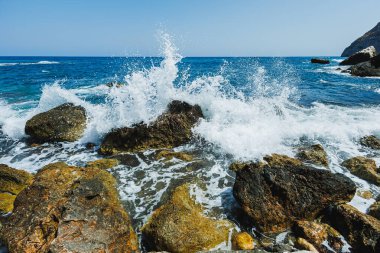 Parlak mavi sular yıpranmış kayalara çarpar, berrak gökyüzünün altında köpüklü sıçramalar yaratır, doğanın gücünü ve dinginliğini yakalar..