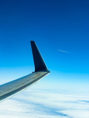 Uçak kanadı kabarık bulutların üzerine uzanırken, uzaktaki başka bir uçağı işaret eden ince beyaz bir iz, parlak mavi gökyüzünün altında sakin bir sahne ortaya çıkıyor..