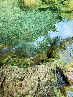 Berrak su, yeşilliklerle çevrili sakin bir bölgede kayaların üzerinde yavaşça süzülür. Güneş ışığı yüzeyden yansıyarak huzurlu manzarayı güçlendirir..