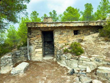 Bu sahnede uzun ağaçların arasına yerleşmiş taştan bir barınak var. Yapıda yaş ve yıpranma izleri var. Girişe giden toprak bir yol var..
