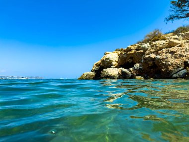 Parlak güneş berrak turkuaz suyun üzerinde parlıyor. Kıyı boyunca kayalık oluşumlara çarpıyor. Huzurlu ve davetkar bir sahil manzarası yaratıyor..