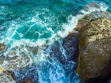 Kristal berraklığında turkuaz sular, engebeli kayalarla buluşurken köpük ve kabararak, güneşli bir sahil gününde doğanın güzelliğinin büyüleyici bir manzarasını yaratır..