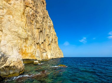 Çarpıcı kıyı manzarası, parlak güneşi ve berrak gökyüzünü yansıtan canlı turkuaz sulardan keskin bir şekilde yükselen yüksek ve engebeli bir uçurumu yansıtıyor, barışçıl bir atmosfer yaratıyor..
