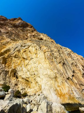 Görüntü, kıyı şeridinden yükselen sarp, renkli kayalıkları yakalıyor, gündüz vakti açık mavi bir gökyüzüne vuran parlak güneş ışığıyla aydınlanıyor..