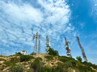 Antenli uzun iletişim kuleleri açık bir günde bulutlarla benekli açık mavi gökyüzünün altında yeşilliklerle çevrili kayalık arazinin üzerinde yükseliyor..