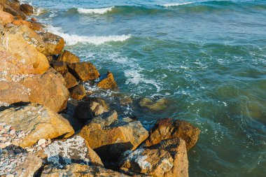 Turkuaz dalgalar ritmik bir şekilde yuvarlanarak, güneşli taşlarla süslenmiş kayalık sahili öperler. Sahil şeridinde doğanın kucakladığı huzurlu bir an..