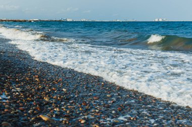 Yumuşak dalgalar, pürüzsüz bir çakıl taşı kumsalında kırılır ve okyanus, gemilerin görülebileceği uzak bir ufka doğru uzanır. Gökyüzü berrak ve berrak, huzurlu atmosferi güçlendiriyor..