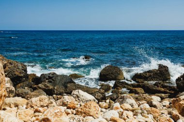 Parlak güneş ışığı dalgaların büyük taşlara çarptığı kayalık bir kıyıda parlar. Açık mavi su ufuk çizgisine kadar uzanıyor ve sakin bir atmosfer yaratıyor..