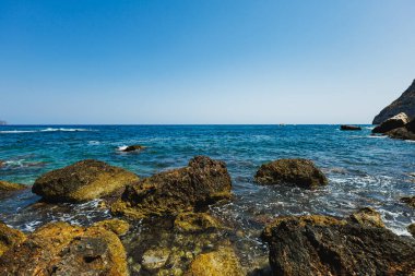Parlak güneş parıldayan denizin üzerinde parlarken dalgalar yavaşça kayalık kıyılara çarpıyor. Uzakta sürüklenen küçük bir tekne, huzurlu manzarayı güzelleştirir..