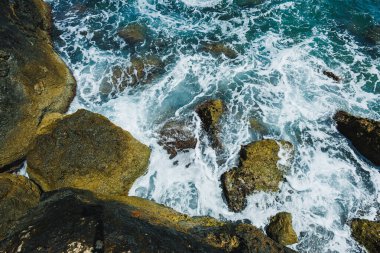 Turkuaz dalgalar açık mavi gökyüzünün altında karanlık ve pürüzlü kayalara çarpıyor ve kıyı şeridinde dinamik bir manzara yaratıyor. Doğanın güzelliği her sıçrayışında büyülenir..