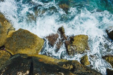 Köpüklü dalgalar, dalgalı bir kıyı şeridi boyunca kayalara çarpar, güneşli bir öğleden sonra parlak mavi gökyüzünü yansıtarak, doğanın deniz kenarındaki güzelliğinin özünü yakalar..