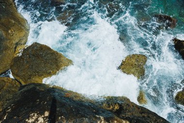 Güçlü dalgalar kıyı kenarındaki sivri kayalara çarpıyor ve parlak öğle güneşinde güzel bir köpük ve sprey gösterisi yaratıyor. Su berrak, mavinin canlı tonlarını sergiliyor..