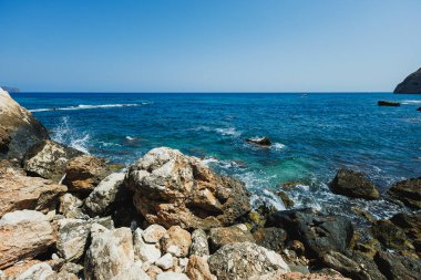 Dalgalar, açık mavi gökyüzünün altında parıldayan kayalık kıyılara hafifçe çarpar. Huzurlu sahne rahatlamaya ve derin düşüncelere davet eder, doğa severler için mükemmeldir..