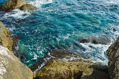 Hafif dalgalar engebeli kayalara çarparken güneş ışığı berrak mavi sularda dans eder ve sıcak bir yaz öğleden sonrası boyunca manzaralı sahil şeridi boyunca rahatlatıcı bir atmosfer yaratır..
