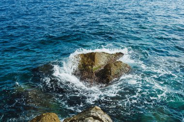 Okyanusun yüzeyinde güneş ışığı parıldıyor dalgalar yavaşça kayalara çarpıyor. Deniz köpüğü gün ortası havasında doğanın ahenginin güzelliğini göstererek yükselir ve dans eder..