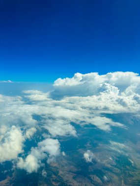 Yumuşak, beyaz bulutlar, yüksek irtifada güneşli bir öğleden sonra uçuşu sırasında bir uçaktan görülen açık mavi gökyüzünün altındaki manzarayı örtüyor..