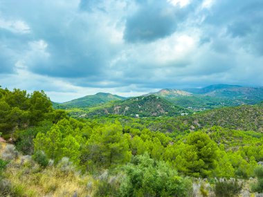Yeşil yemyeşil tepeler, ağaçlarla benekli arazi boyunca uzanırken, dramatik bulutlar gökyüzünde beliriyor. Doğa bu güzel sahneye huzur ve sükunet katar..