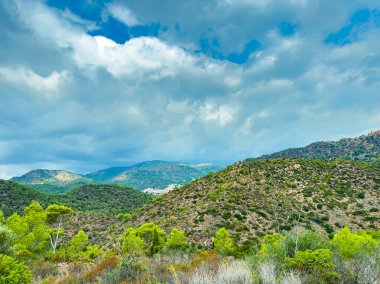 Canlı yeşil tepeler, parçalı bulutlu bir gökyüzünün altında, manzara boyunca uzanıyor ve sakin bir atmosfer yaratıyor. Bu dingin ortam, doğanın güzelliğini keşfetmeye ya da gevşetmeye davet eder..