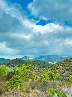Parlak yeşilliklerle kaplı yuvarlanan tepeler bulutlarla dolu bir gökyüzünün altında, öğleden sonra ortasında doğada barışçıl ve dramatik bir sahne yaratıyor..