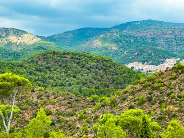 Canlı yeşil tepeler manzara boyunca uzanıyor, ağaçların arasından gözetleyen küçük bir köy, bulutlu bir gökyüzünün altında sakin bir manzara yaratıyor..