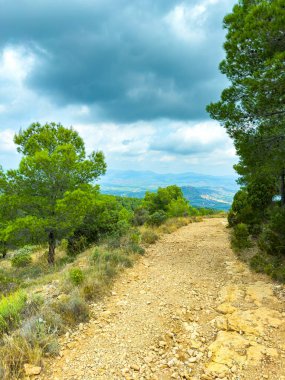 Dolambaçlı bir toprak yol gezginleri hareketli yeşil ağaçların arasından kasvetli bir gökyüzünün altına davet eder. Dağlar arka planda uzanıyor, doğaya huzurlu bir kaçış yaratıyor..